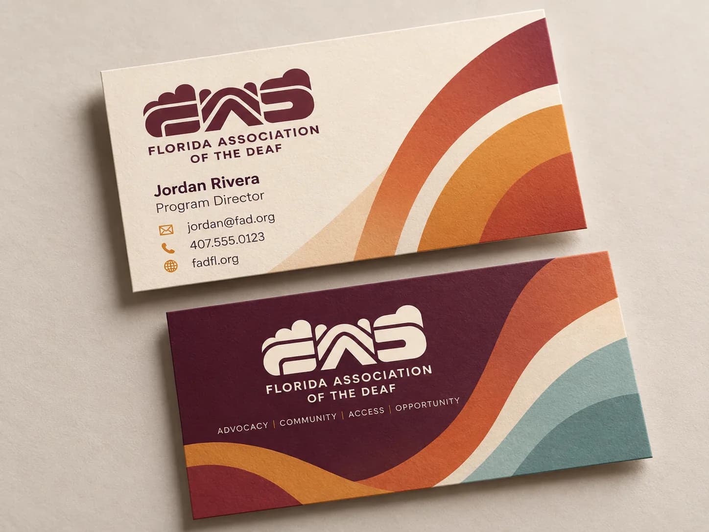 Two Sunrise business cards on a linen surface: a cream front with the plum rounded FAD wordmark and contact details beside sweeping orange, marigold, and plum arcs, and a plum back with the wordmark in cream above Advocacy, Community, Access, Opportunity, over orange, cream, and teal waves.